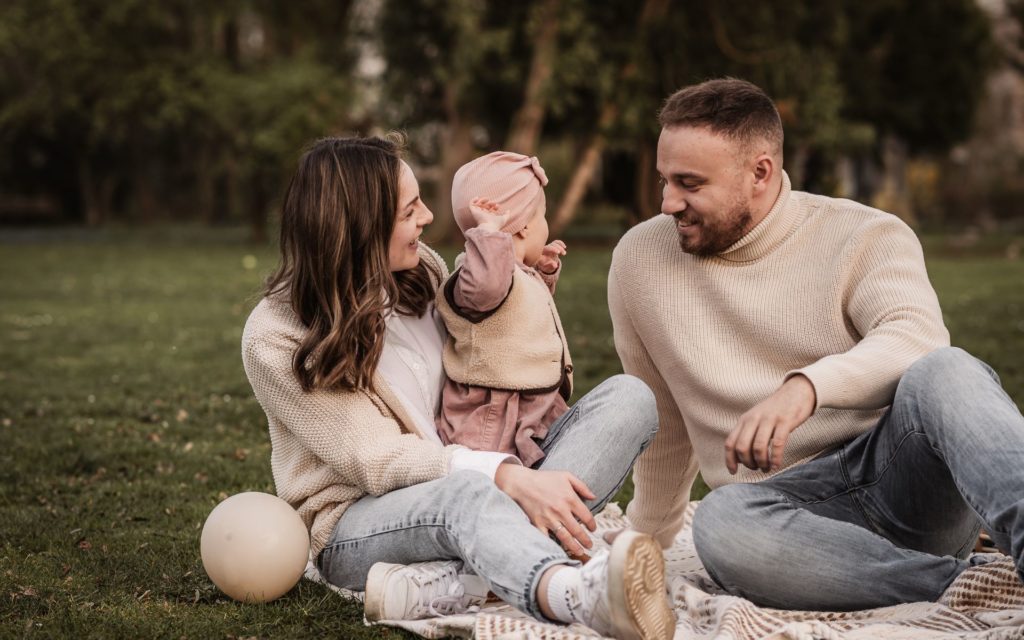 Mama, Papa und Kleinkind im Park auf einer Picknickdecke. Sie lachen miteinander und spielen mit Luftballons.