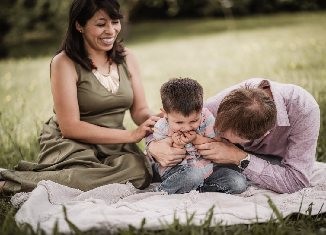 Familien mit Mama, Papa und Sohn gemütlich auf einer Picknickdecke im Freien. Papa und Sohn albern zusammen herum.