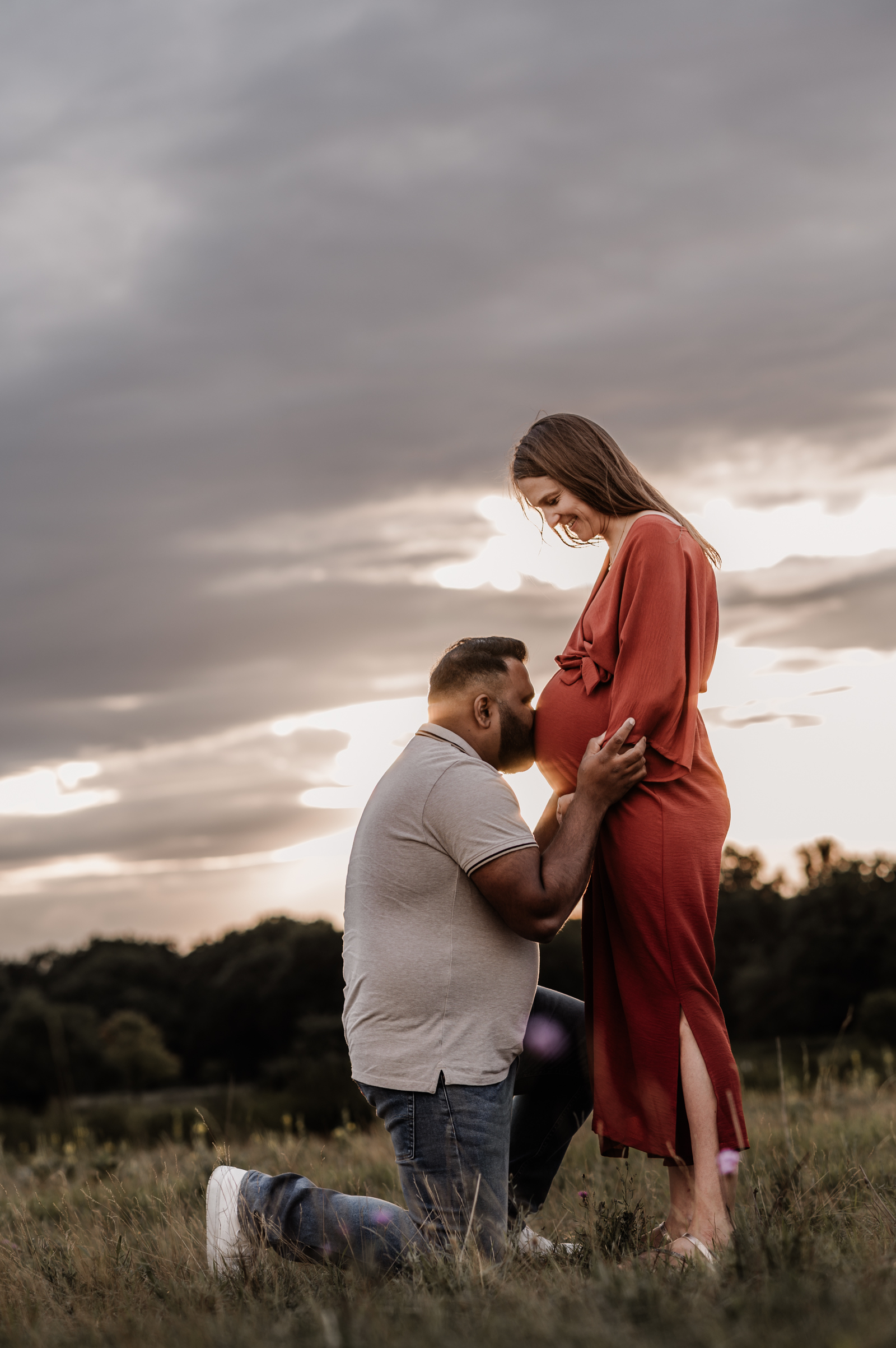 Werdende Eltern bei Sonnenuntergang in der Natur. Papa kniet vor der werdenden Mama und gibt Babybauch einen Kuss.