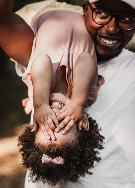 Familienfoto mit Kleinkind und Daddy. Das Mädchen hält nich die Augen zu, während ihr Papa es über die Schulter gelegt trägt. Das Mädchen und Papa lachen gemeinsam und haben Spaß im Freien.