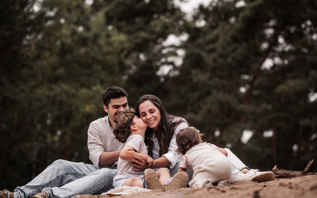 Eine Familie mit Mama, Papa und zwei Kindern auf einer Sanddüne vor waldigem Hintergrund. Sie strahlen und verbringen gemeinsame Zeit bei einem Familienfotoshooting in Nürnberg.