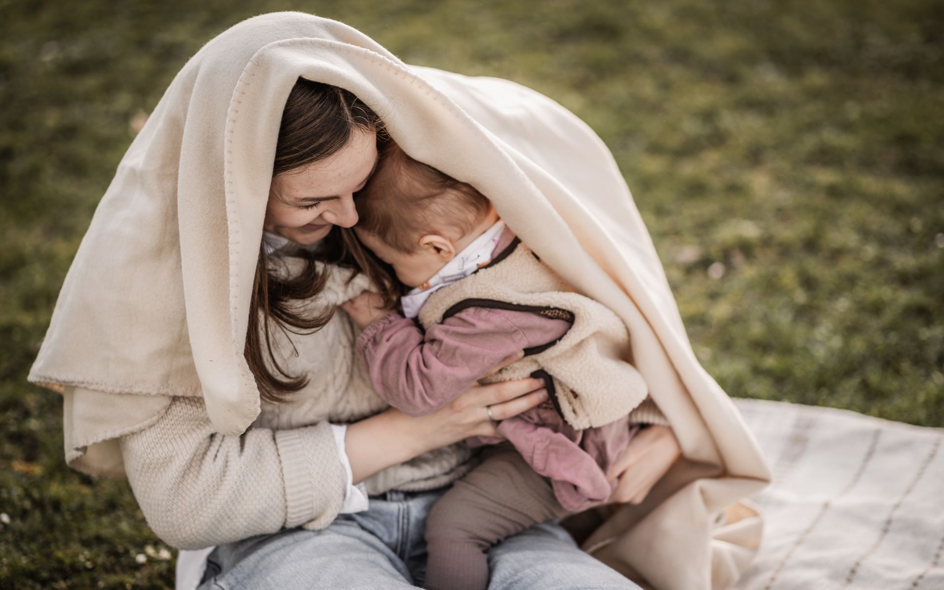 Mama mit Kleinkind kuschelt unter einer cremefarbenen Decke und beide kichern dabei.