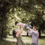 Mama und Papa auf Picknickdecke im Grünen heben ihren Sohne in die Höhe und feiern seinen Geburtstag. Alle kichern und lachen gemainsam.