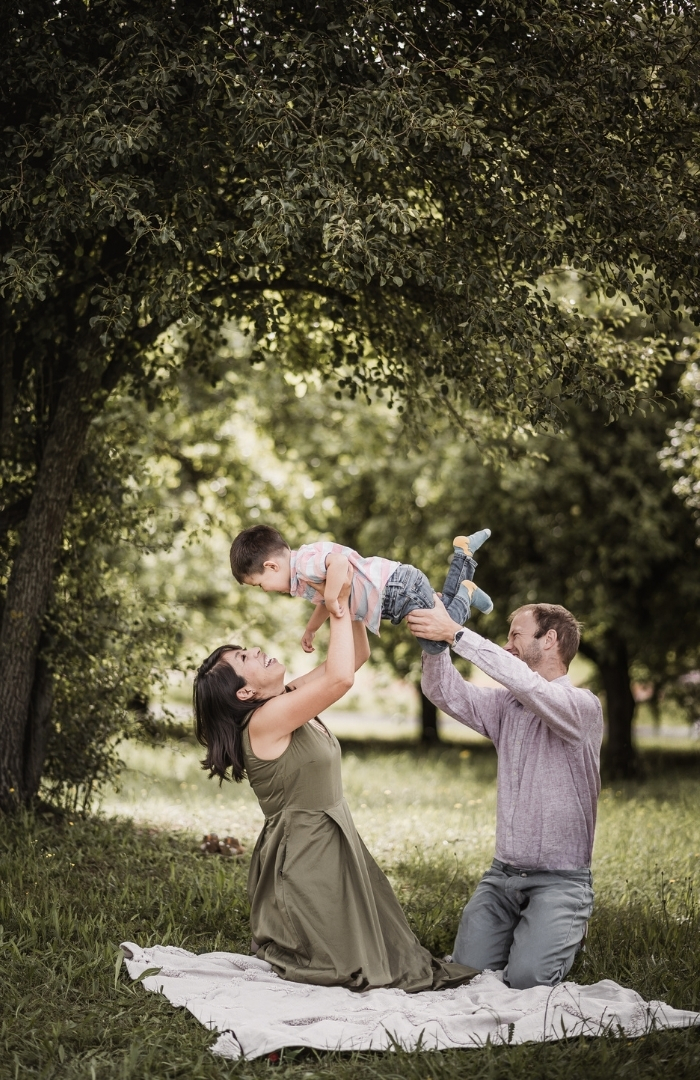 Mama und Papa auf Picknickdecke im Grünen heben ihren Sohne in die Höhe und feiern seinen Geburtstag. Alle kichern und lachen gemainsam.