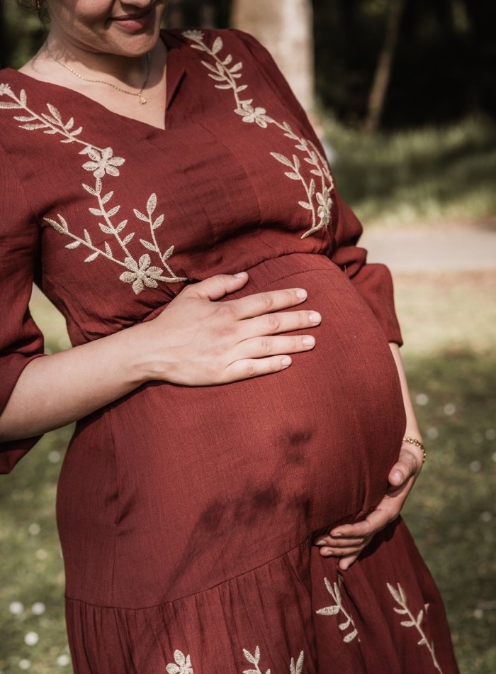 Schwangere Mama in Fürth in der Natur. Liebevoll streichelt sie sich über den Babybauch. Die trägt ein rostrotes Kleid und strahlt richtig.