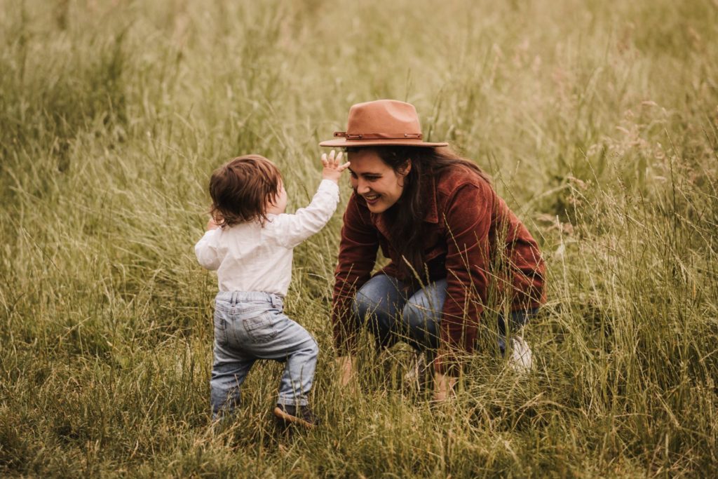 Mama mit ihrem kleinen Jungen in einer schönen Wiese. Er läuft auf sie zu und versucht den Hut vom Kopf zu nehmen, während sie lachend vor ihm hockt.