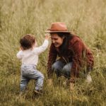 Mama mit ihrem kleinen Jungen in einer schönen Wiese. Er läuft auf sie zu und versucht den Hut vom Kopf zu nehmen, während sie lachend vor ihm hockt.
