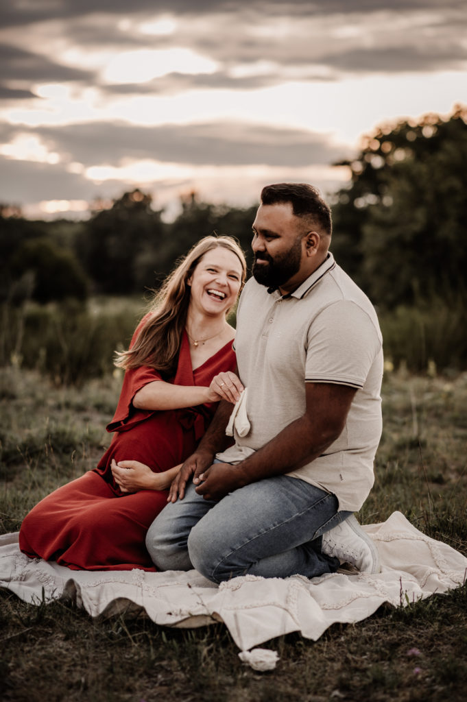 Paar beim Babybauchshooting in der Natur in Nürnberg. Sie sitzen lachend auf einer Decke mit kleinen Babysöckchen in der Hand.