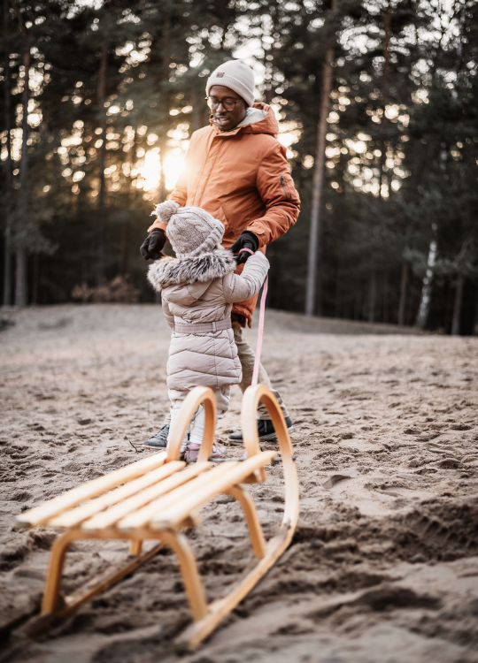 Kleinkind mit Daddy und Schlitten auf einer Sanddüne bei untergehender Sonne umrahmt von einem Kiefernwald