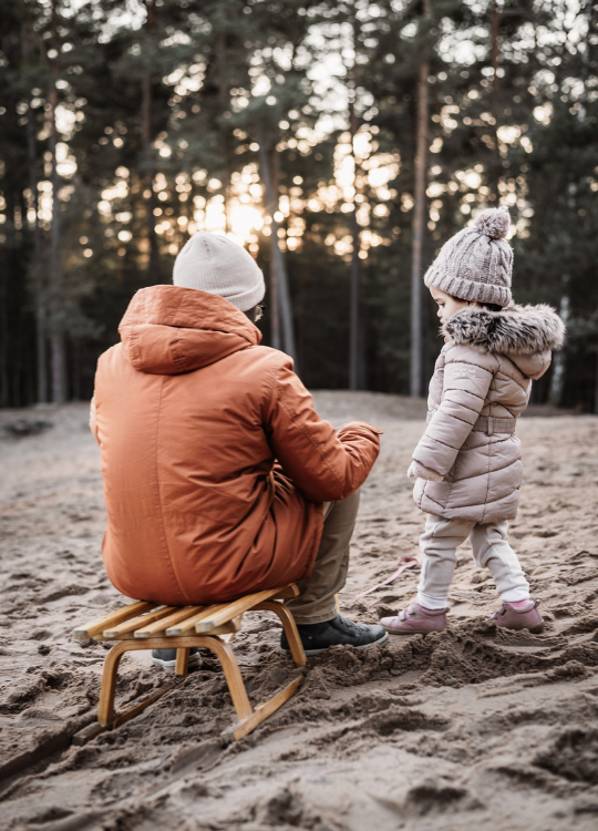 Kleinkind mit Daddy und Schlitten auf einer Sanddüne bei untergehender Sonne umrahmt von einem Kiefernwald