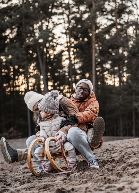 Kleinkind mit Mama und Papa auf Schlitten auf einer Sanddüne bei untergehender Sonne umrahmt von einem Kiefernwald