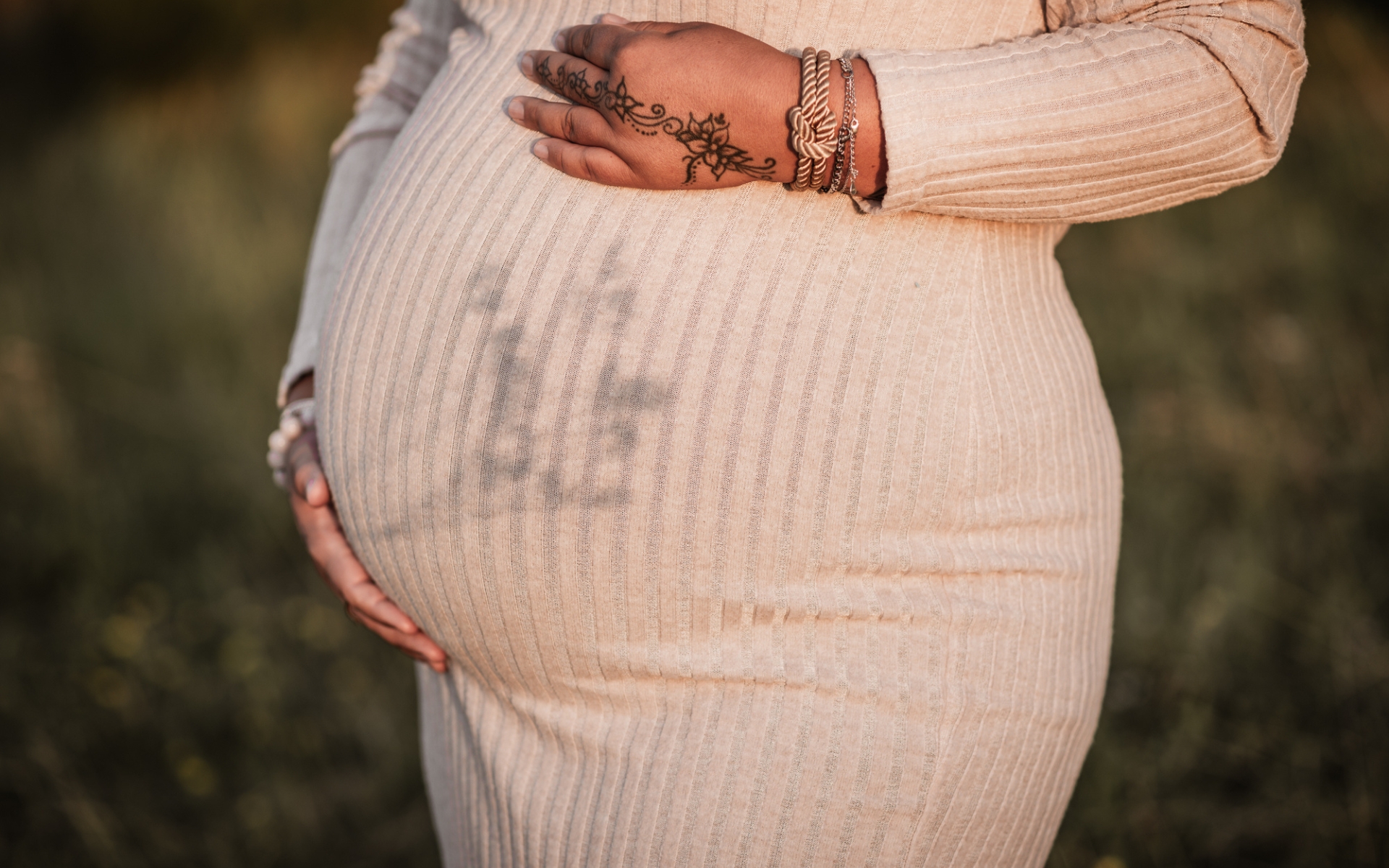 Nahaufnahme einer werdenden Mama in cremefarbenden Strickkleid. Die untergehende Sonne wirft ein Schattenspiel auf ihren Babybauch.