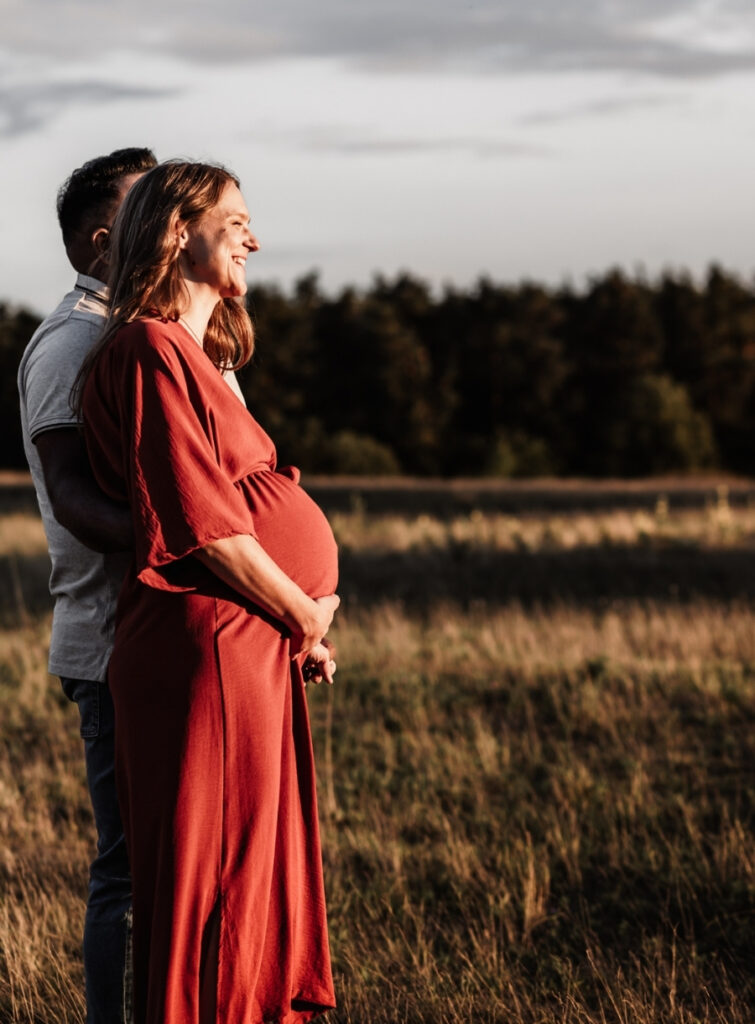 Paar beim Babybauchshooting im Sonnenuntergangslicht auf einer offenen Wiesenfläche mit Wald im Hintergrund