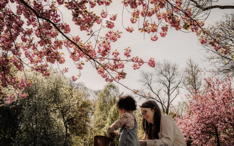Mama mit ihrer Tochter, welche ein Laufrauf in der Hand hält. Sie sind unscharf im Vordergrund vor rosa blühenden Bäumen.