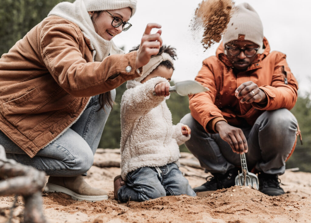 Mama mit Kleinkind und Papa spielen im Sand. Das kleine Mädchen wirft sand um sich