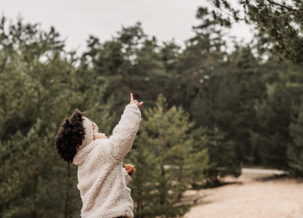 Kleines Mädchen mit Afrolocken und weißen Stirnband auf einem sandigen Weg inmitten von Wald zeigt nach oben mit dem Zeigefinger auf etwas, dass sie entdeckt hat