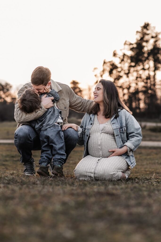 Familie mit Kleinkind und Babybauch bei Sonnenuntergang. Sie lachen und kuscheln auf einer Wiese während die Sonne im Hintergrund untergeht.