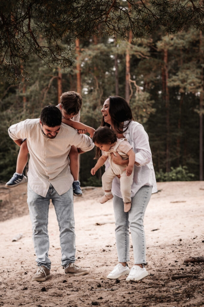 Familie mit zwei Kindern auf dem Arm lacht herzlich unter Kieferbäumen auf der Sanddüne in Nürnberg