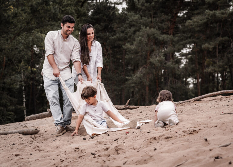 Familie mit zwei Kindern auf der Sanddüne bei Nürnberg ziehen ihr Kind mit Picknickdecke durch den Sand und lachen zusammen, während ein Kleindkind im Sand krabbelt.