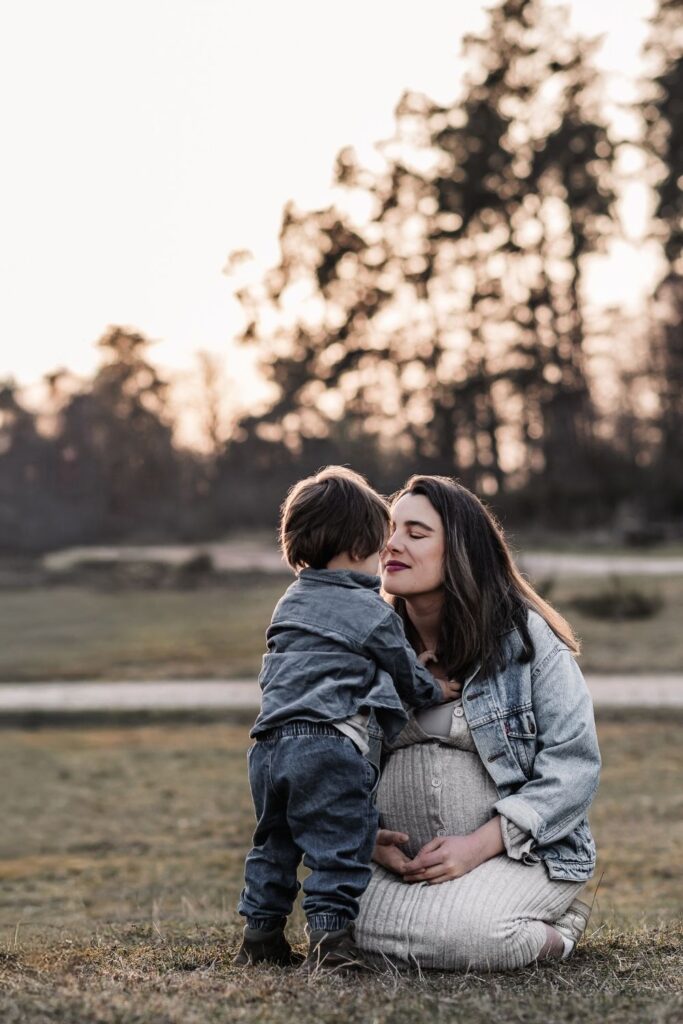 Schwangere Mama mit ihrem Sohn. Sie kniet auf der Wiese während ihr Sohn ihr eine Umarmung gibt.