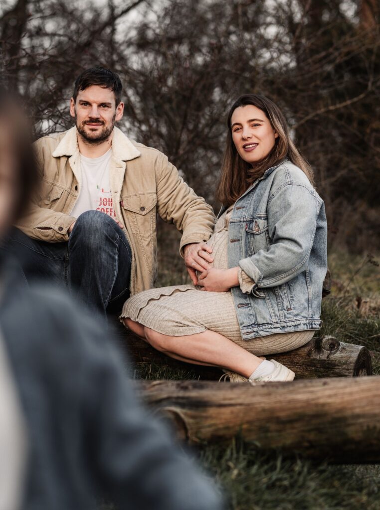 Papa und Mama sitzen auf einem Baumstamm. Die Frau ist schwanger. Papa streichelt zart über den Bauch. Im Vordergrund ist ihr zweijähriger Sohn unscharf zu sehen, der auf die Kamera zurennt.