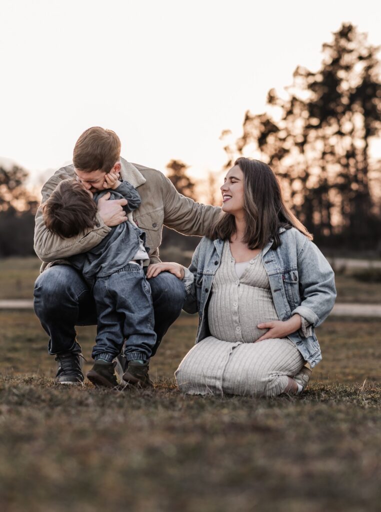 Familien draußen in der Natur beim Familien und Babybauchshooting. Sie genießen die gemeinsame Zeit auf einer Wiese umgeben von Wald. Papa kuschelt seinen Sohn während schwangere Mama daneben in der Wiese kniet und lächelnd zuschaut.