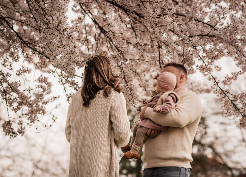 Familie unter einem rosa blühenden Baum. Sie schauen hinauf zu den Blüten und bewundern diese. Das Foto ist von hinten aufgenommen.