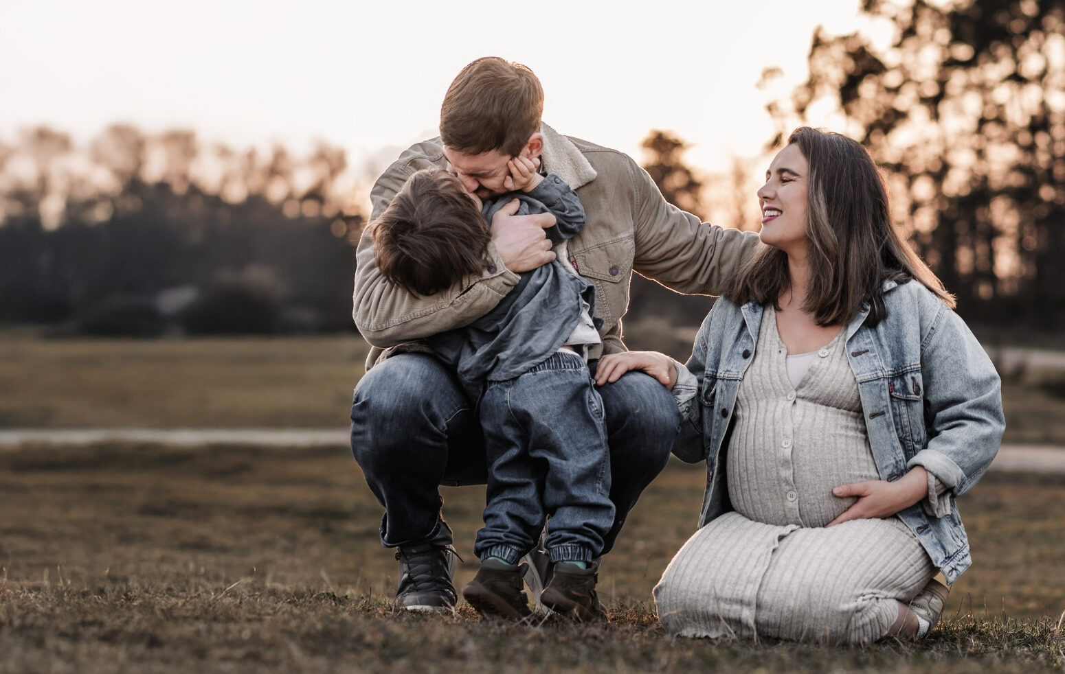 Familie mit kleinem Kind beim Babybauch Shooting in der Natur in Nuernberg und Fürth. Sie lachen gemeinsam und knien auf einer Wiese beim Sonnenuntergang.