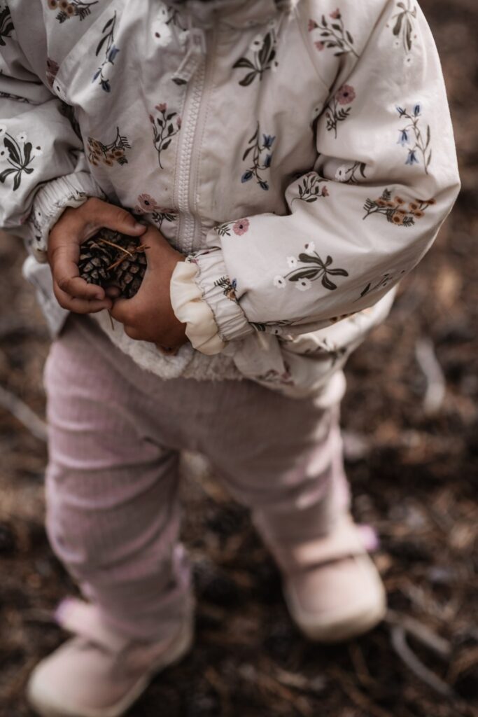Ein kleines Mädchen läuft auf sandigem weg. Sie hat einige Kiefernzapfen in der Hand. Das Foto zeigt sie von oben.