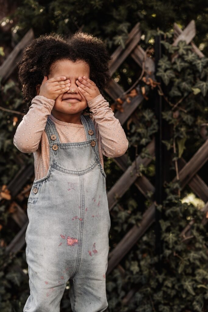 Kleines Mädchen in Jeans-Overall versteckt sich hinter ihren Händen. Sie steht draußen in der Natur vor einem Holzzaun.
