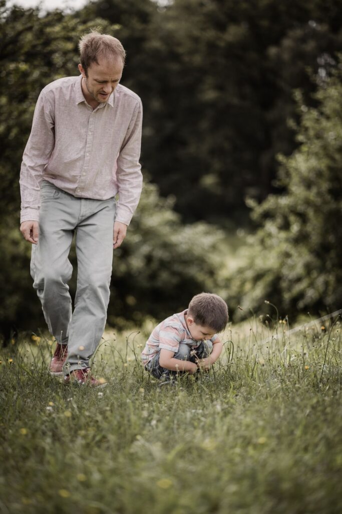 Papa geht beim Familienshooting mit seinem 3-jährigen Sohn über eine Wiese. Der Junge betrachtet etwas im Gras.