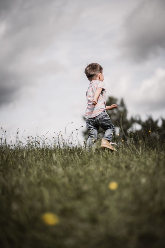 3-jähriger Junge beim Familienshooting rennt über eine grüne Wiese mit Löwenzahl-Blumen weg von der Kamera.