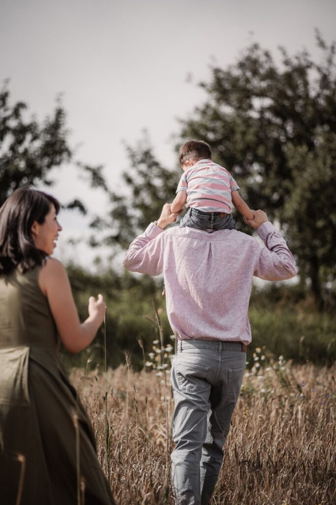 Familienfoto mit Kleinkind. Mama schaut zu, wie Papa ihren Sohn auf den Schultern von durch ein Getreidefeld trägt.