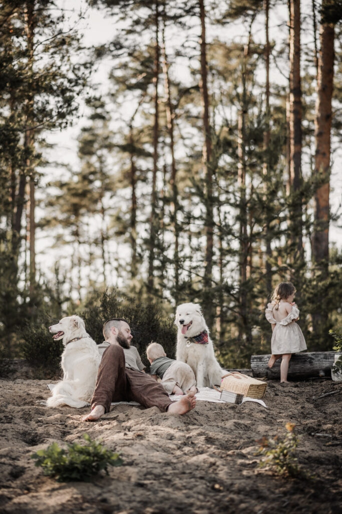 Papa liegt gemütlich auf einer Sanddüne auf einer Picknickdecke. Er iwrkt entspannt. Sein Sohne (1 Jahr) und 3-jährige Tochter bewegen sich um ihn herum. Ein großer weißer Hund sitzt entspannt daneben.