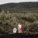 Mama mit großem weißen Hund an der Leine läuft einen sandigen Weg hoch. Im Hintergrund ist viel weiter Wald zu sehen. Ein Kleinkind ist versteckt hinter dem Hund zu sehen.
